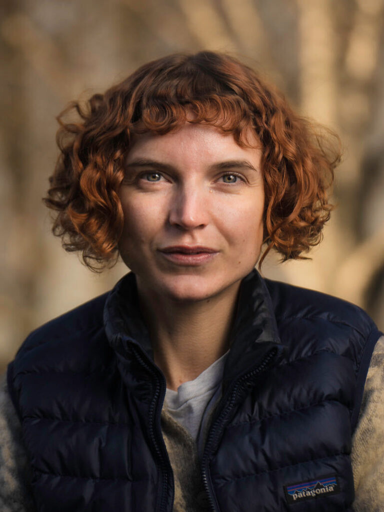 Portrait of a woman with short curly red hair, outdoors, wearing a navy Patagonia quilted vest and light-colored shirt.