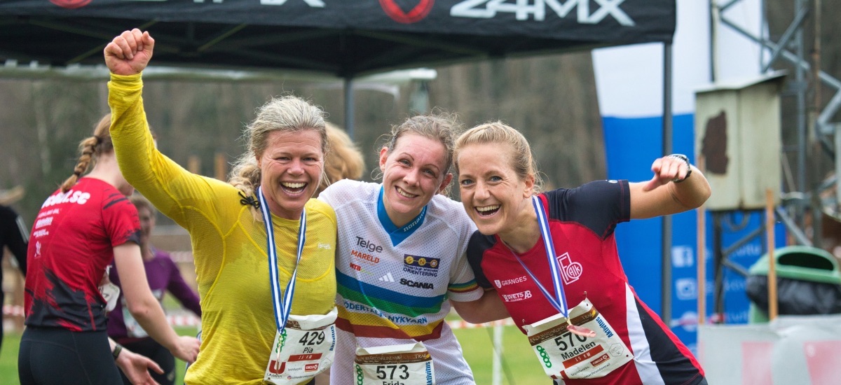 Three female runners wearing bibs and medals celebrate together at a race finish, arms around each other.