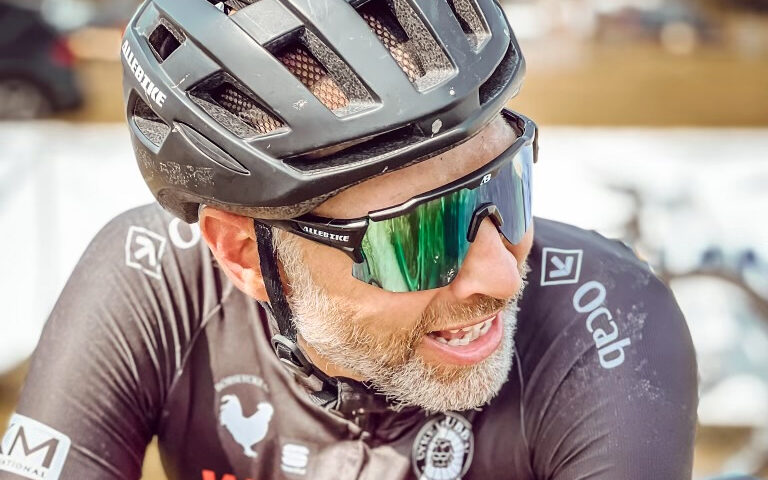 Close-up of a male cyclist wearing a black helmet and reflective green sunglasses, smiling during a ride outdoors.