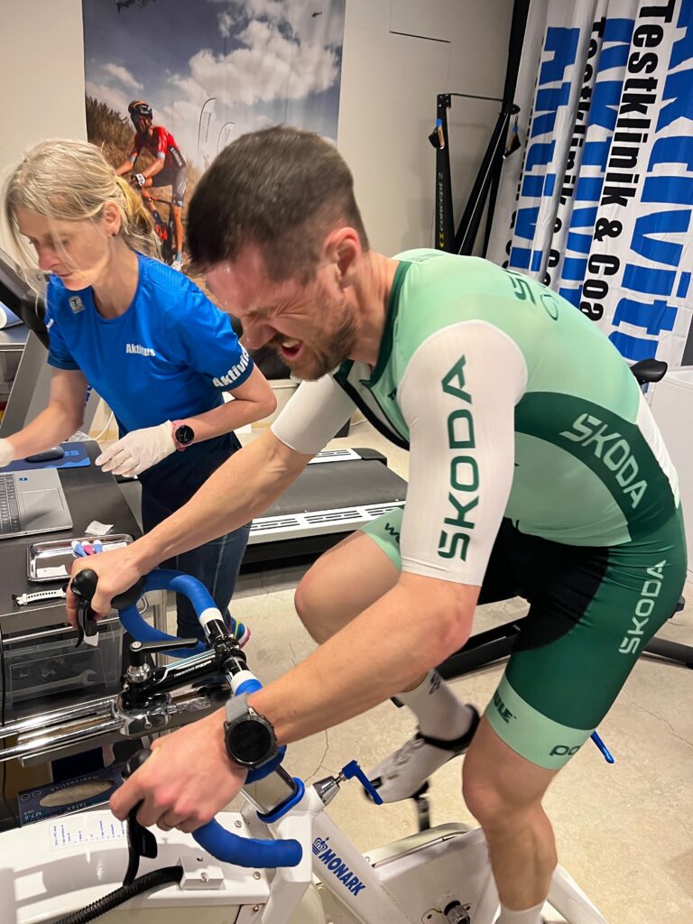 IMG_1346 - Aktivitus Two cyclists on stationary bikes indoors; man in green Skoda kit leans forward as woman in blue Aktivus shirt watches nearby.