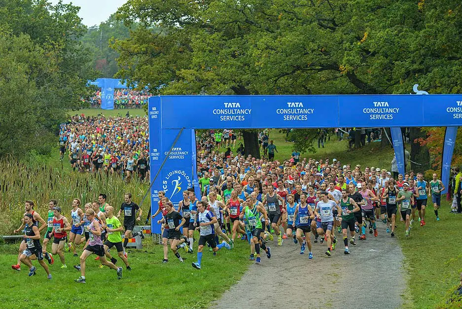 A large group of runners starts a race under a blue sponsor arch labeled Tata Consultancy Services in a park.