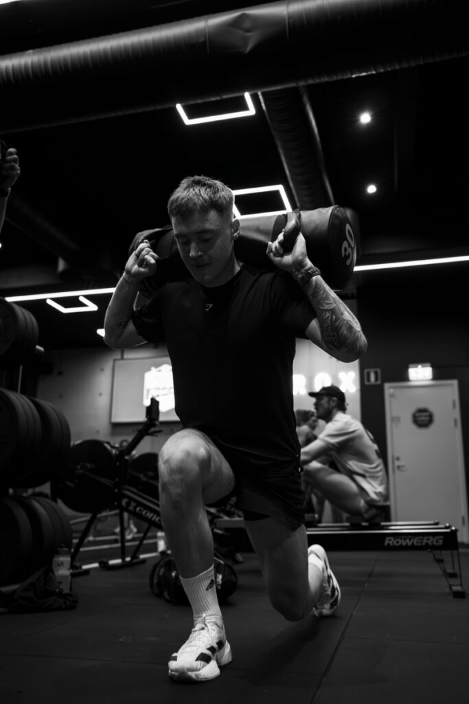 Man in a black shirt lunges forward with a heavy bar across his shoulders in a dim gym setting.