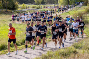 Large group of runners competing in a charity or marathon along a rural dirt path with grassy fields and trees in the background under sunny skies, many wearing bib numbers and athletic gear.