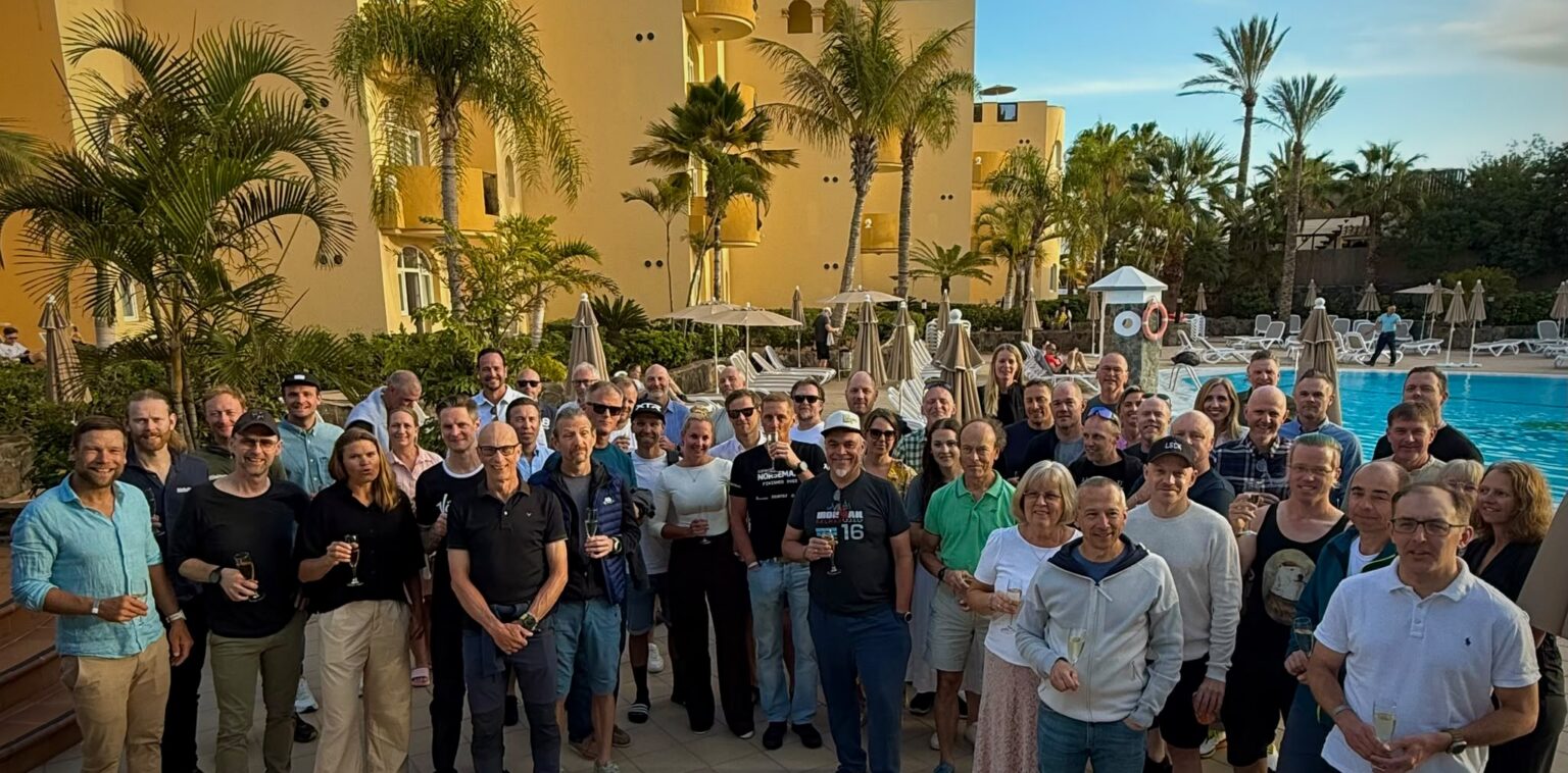 Large group of people posing for a photo by a pool at a sunny resort, with palm trees and yellow buildings in the background.