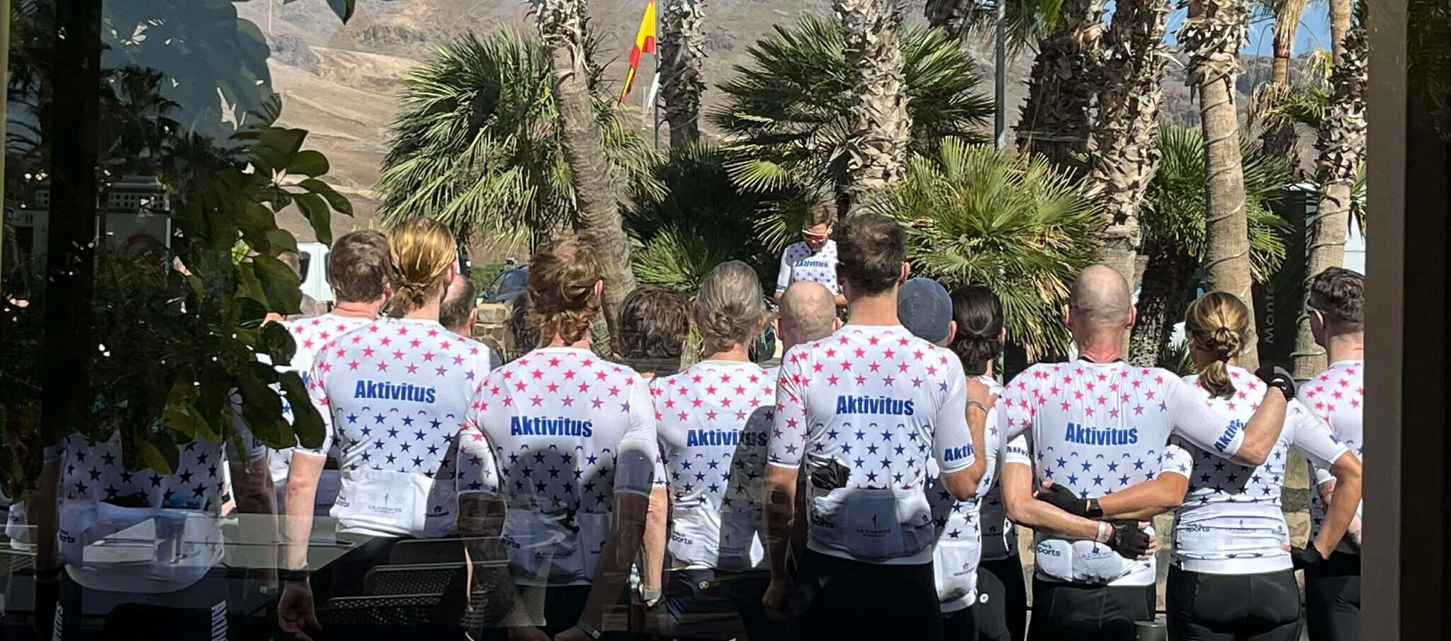 Group of people in white star-pattern shirts with 'Aktivitus' on back, standing outdoors among palm trees and hills.