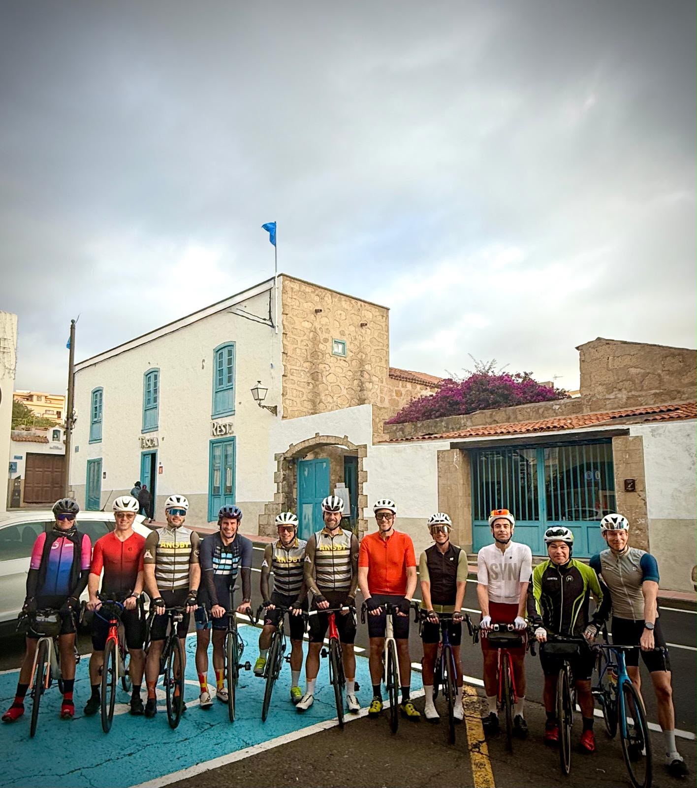 Group of cyclists in colorful jerseys and helmets posing with their bikes on a blue crossing in front of a white building with blue doors and windows nearby.