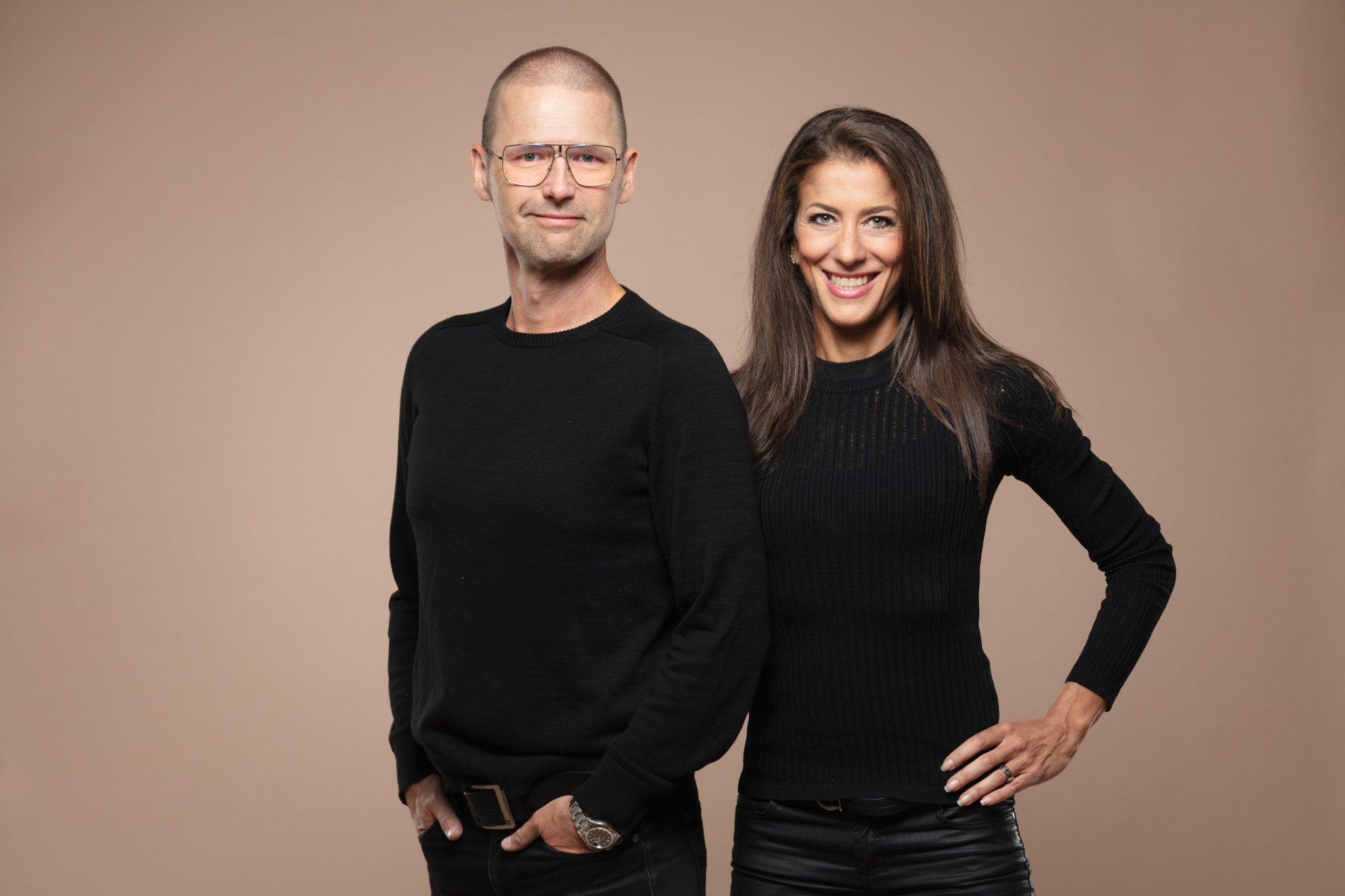 Two adults standing side by side, both in black sweaters, smiling at the camera against a tan backdrop.
