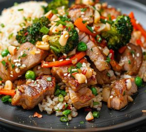 Stir-fried pork with broccoli, bell peppers, green onions, and sesame seeds over rice on a dark plate