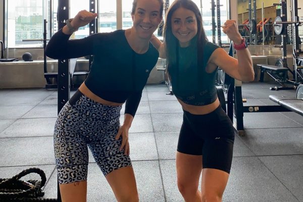 Två kvinnor som ler och poserar med böjda armar i en gymmiljö, iklädda atletiska kläder. Träningsutrustning syns i bakgrunden. Två kvinnor som ler och poserar med böjda armar i en gymmiljö, iklädda atletiska kläder. Träningsutrustning syns i bakgrunden.